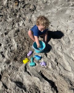 Little blonde boy playing in white sand with a blue bucket that folds down, a yellow crab, purple sea horse and purple turtle sand toy. 